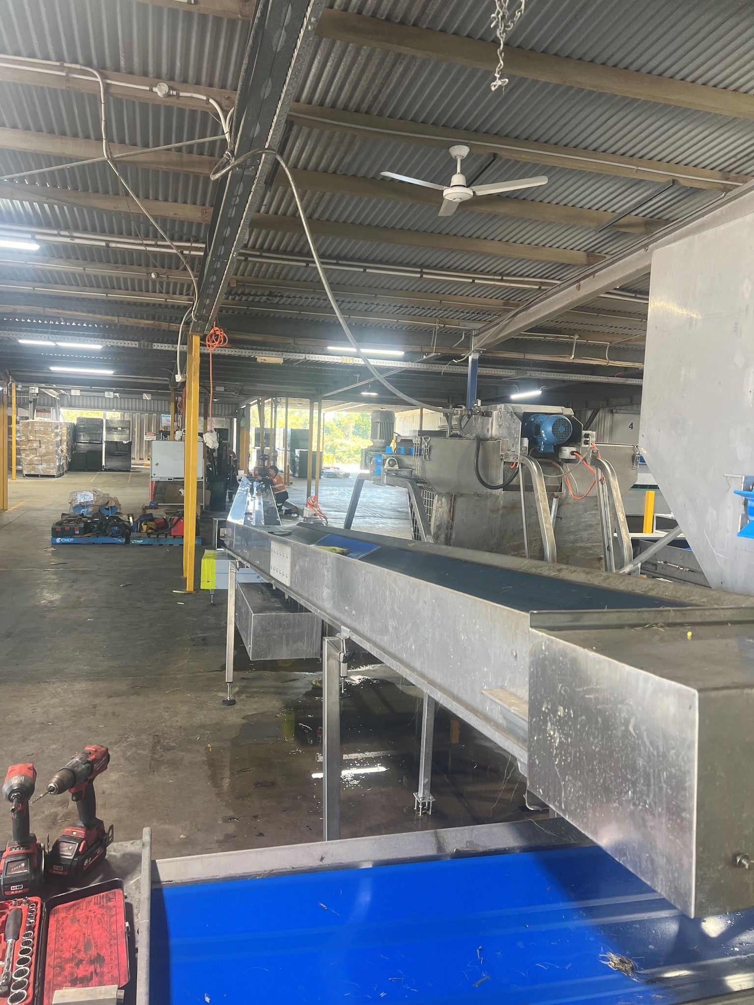 Industrial kitchen with stainless steel prep tables, blue bin, and ceiling fans in an open workspace