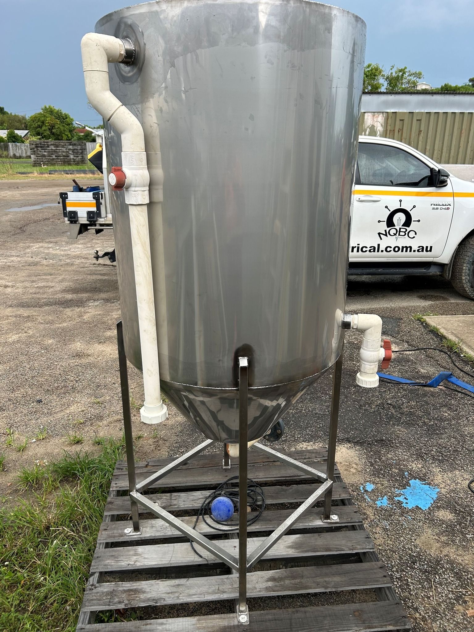 Large silver industrial tank on a pallet in a gravel lot, with pipes and a white utility truck behind it