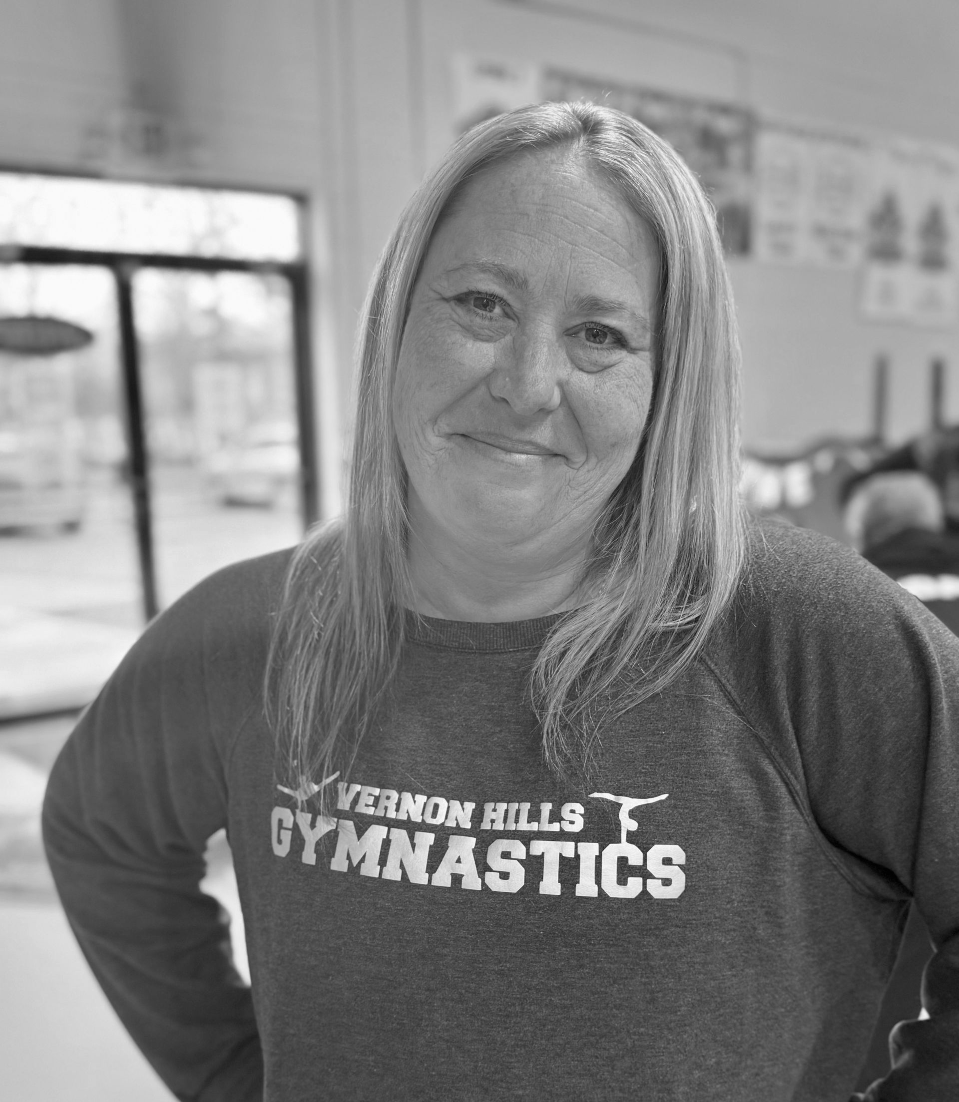 Woman smiling in a gymnastics shirt, standing indoors with hands on hips.