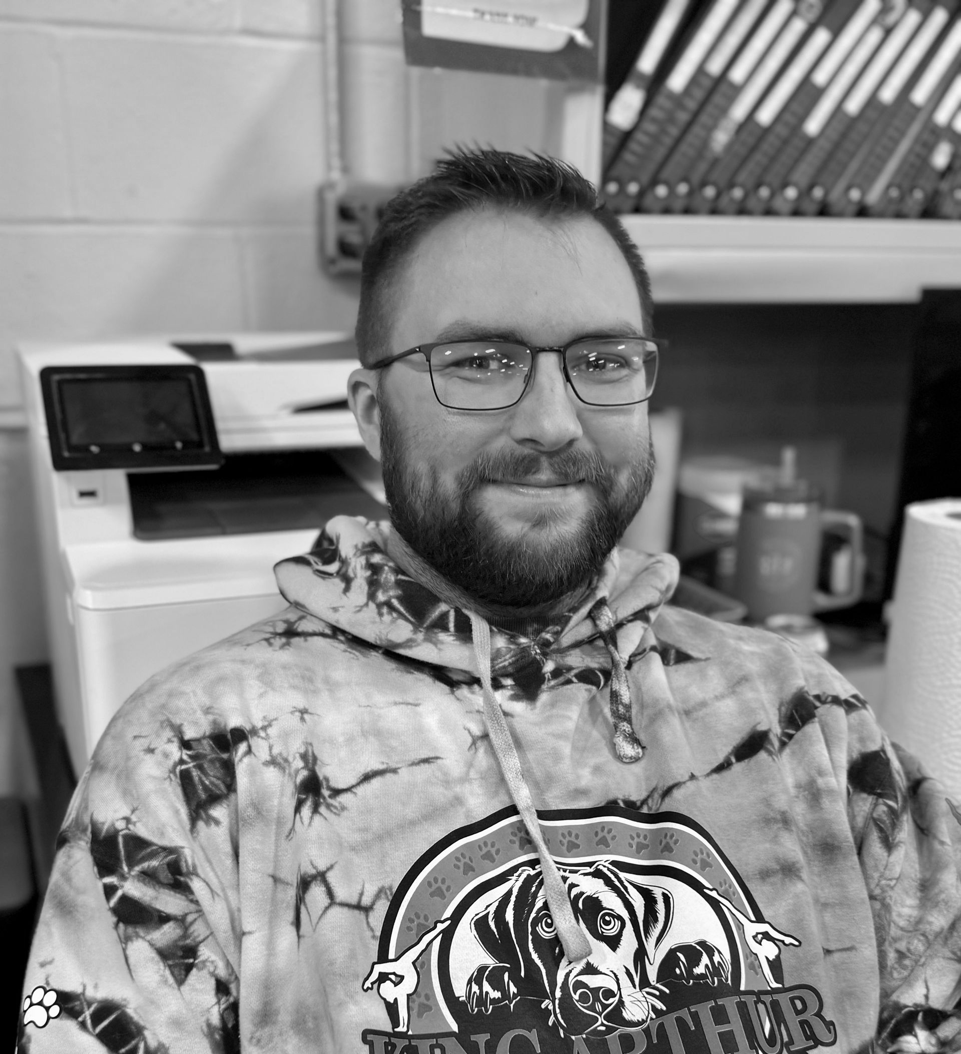 Smiling person in a tie-dye hoodie and glasses seated in a kitchen or break room