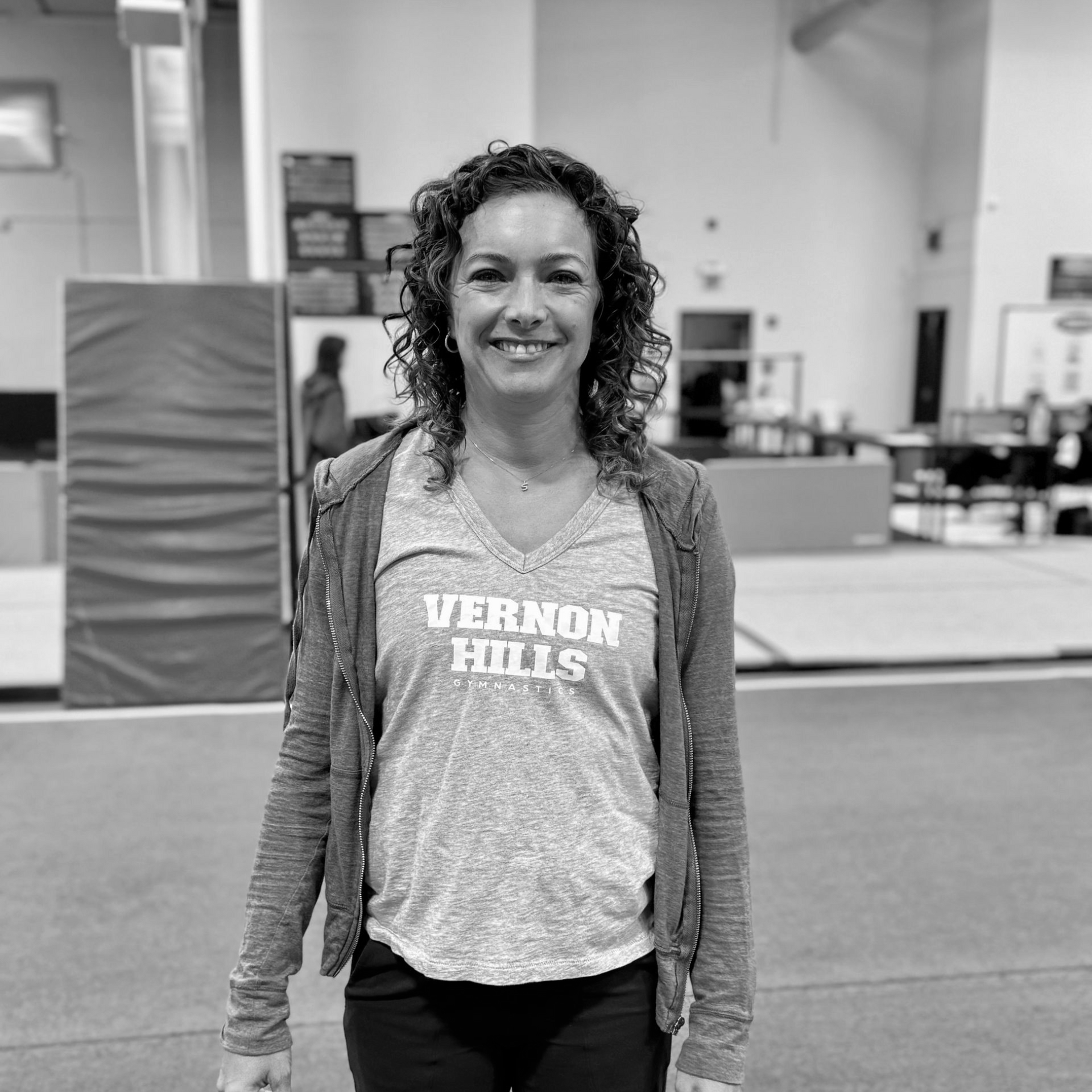 Person smiling in a gym, wearing a Vernon Hills T-shirt and hoodie, standing on a padded floor.