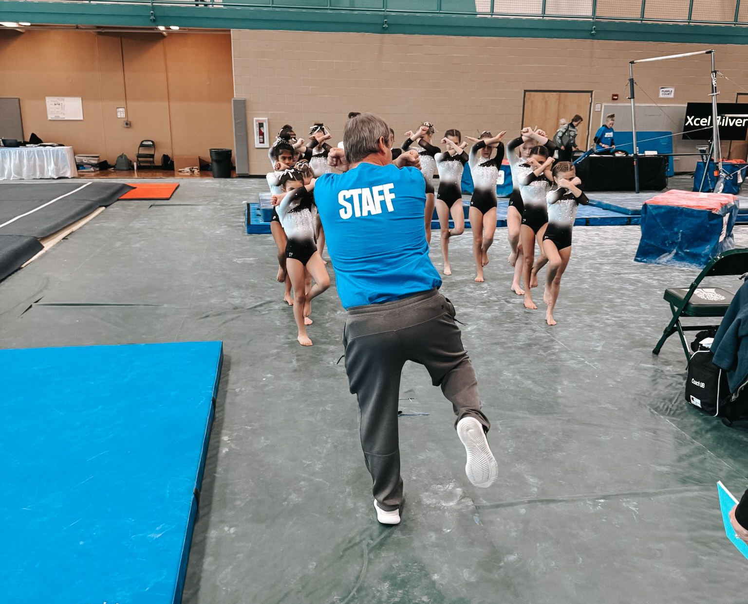 Gymnastics staff member in blue shirt runs behind young gymnasts on a floor mat in a gymnasium