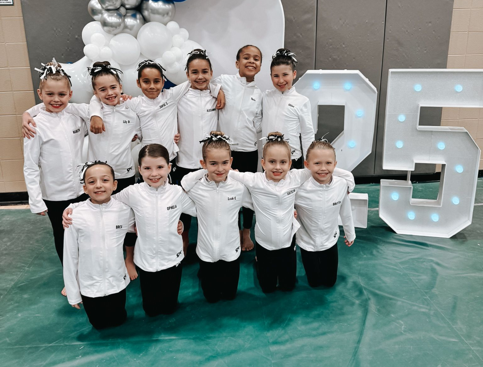 Group of young dancers posing in white outfits in front of large illuminated 25 numbers.