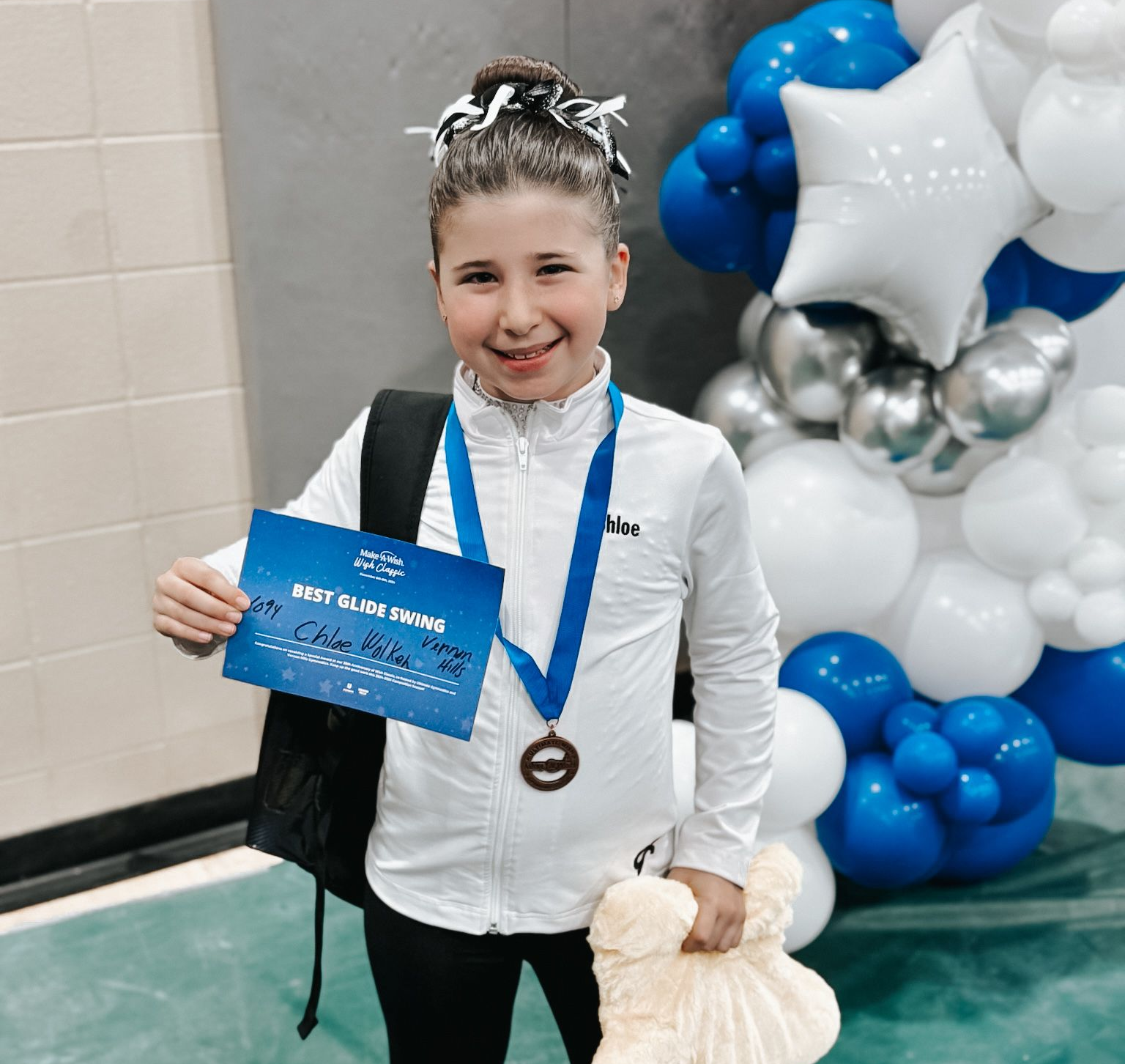 Child holding a blue certificate and medal, standing by blue-and-white balloon decorations.
