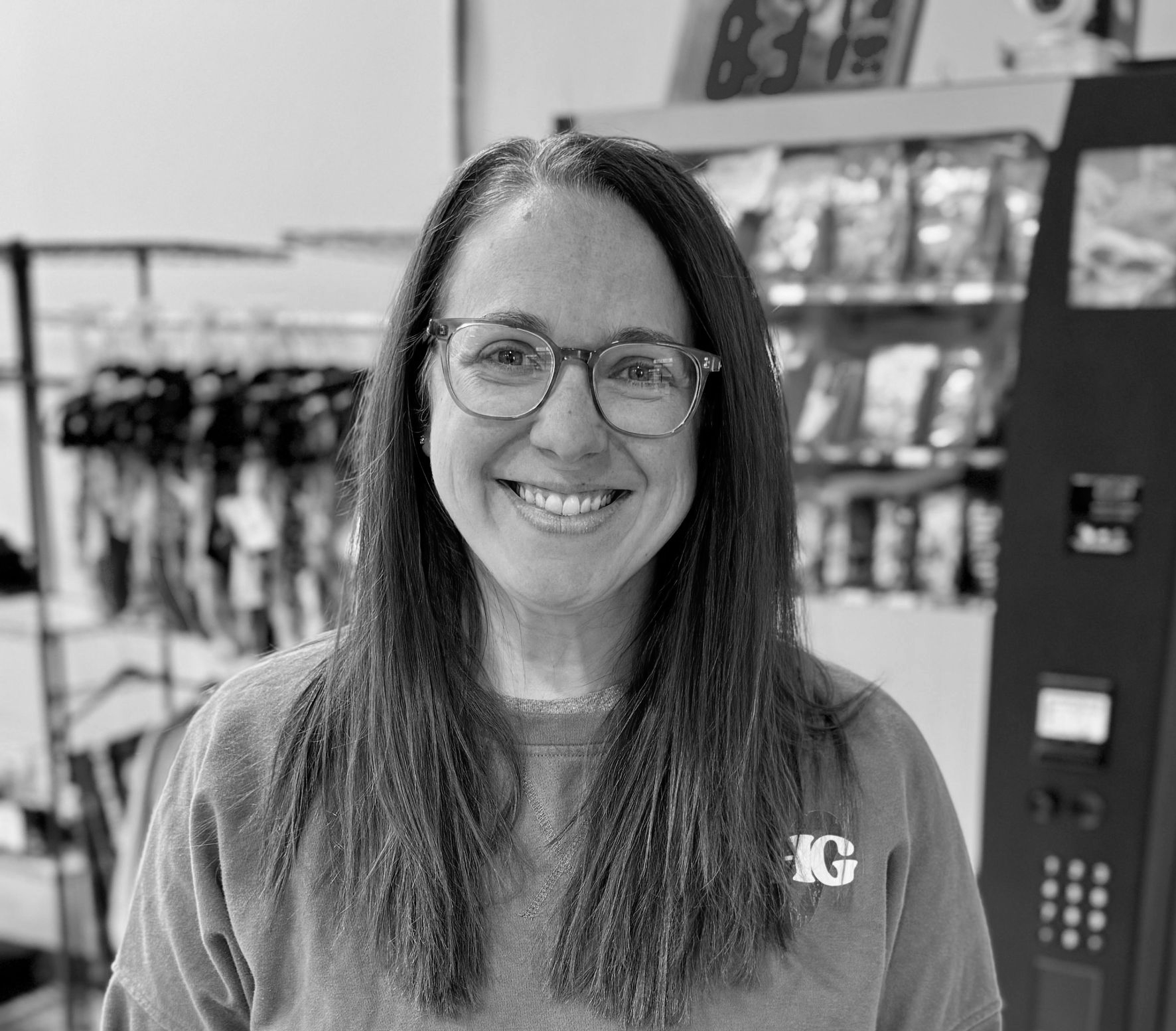 Smiling woman with glasses in a store aisle, standing in front of hanging merchandise.