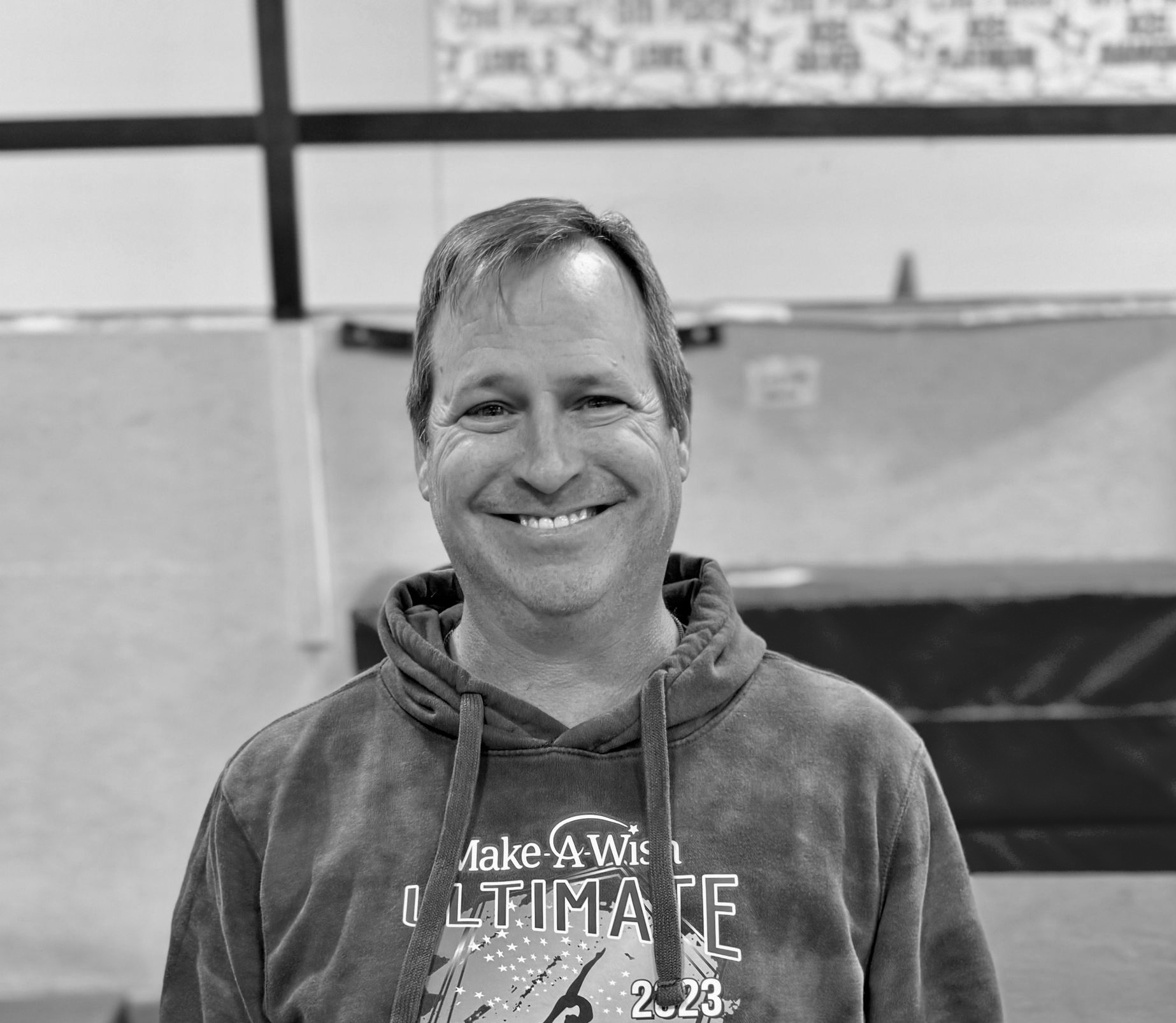 Smiling man in a hoodie standing indoors against a gym wall.