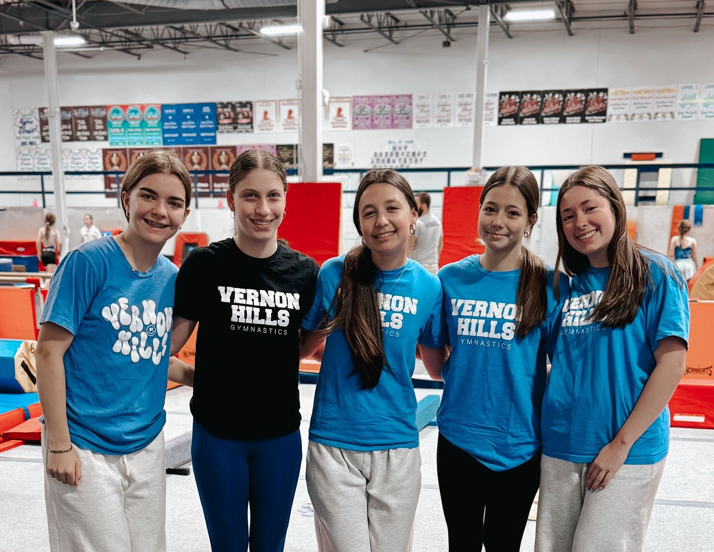 Five people in blue event T-shirts standing together in an indoor sports venue, smiling at the camera.