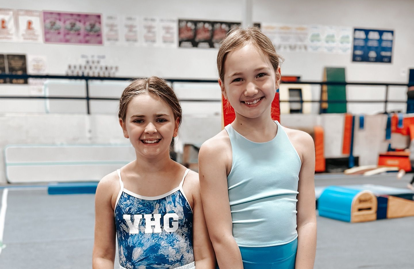 Two smiling gymnasts standing in a gymnastics gym, wearing blue leotards.