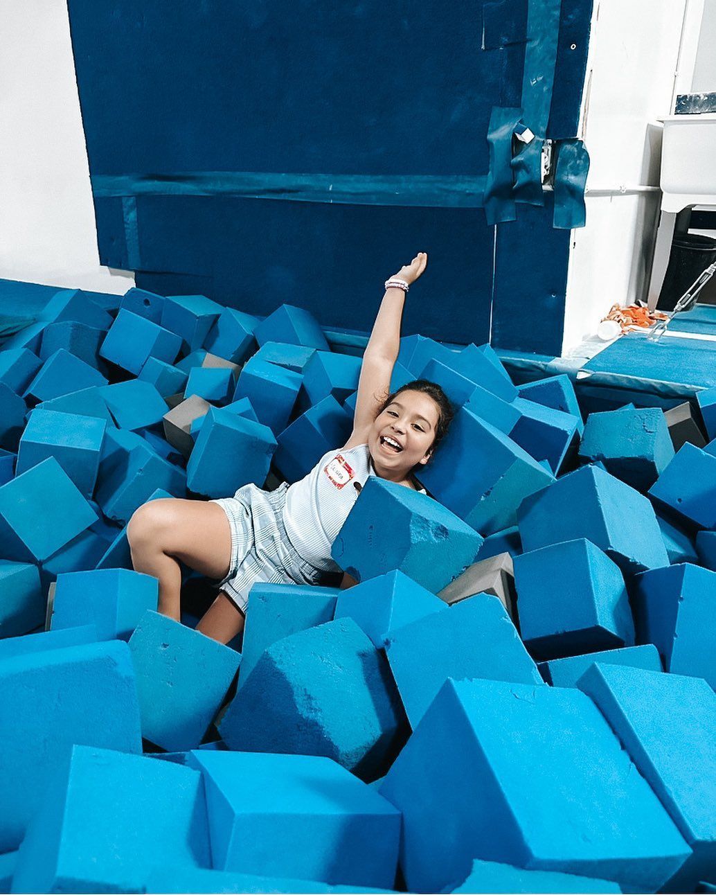 Child smiling in a blue foam pit, raising one arm beside stacked blue blocks
