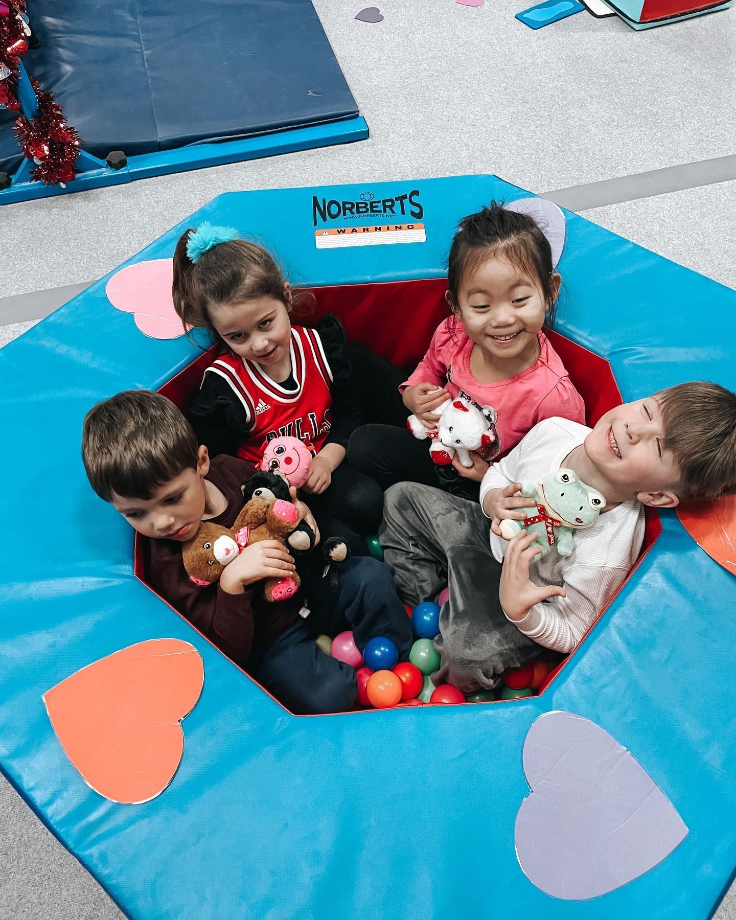 Four children sit in a colorful padded play pit holding stuffed animals.