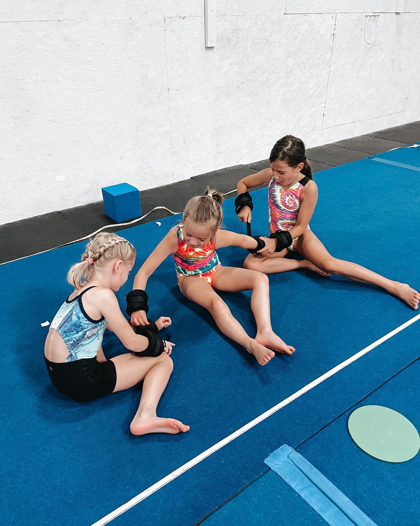 Three children sit on blue gym mats, holding black training pads in a dojo.