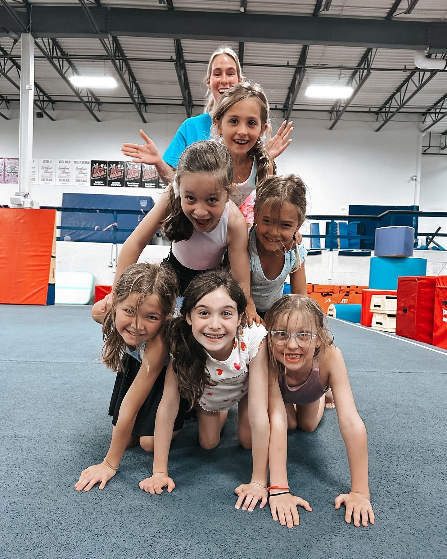 Six children stacked in a human pyramid on a gym mat, smiling in an indoor gymnastics facility.
