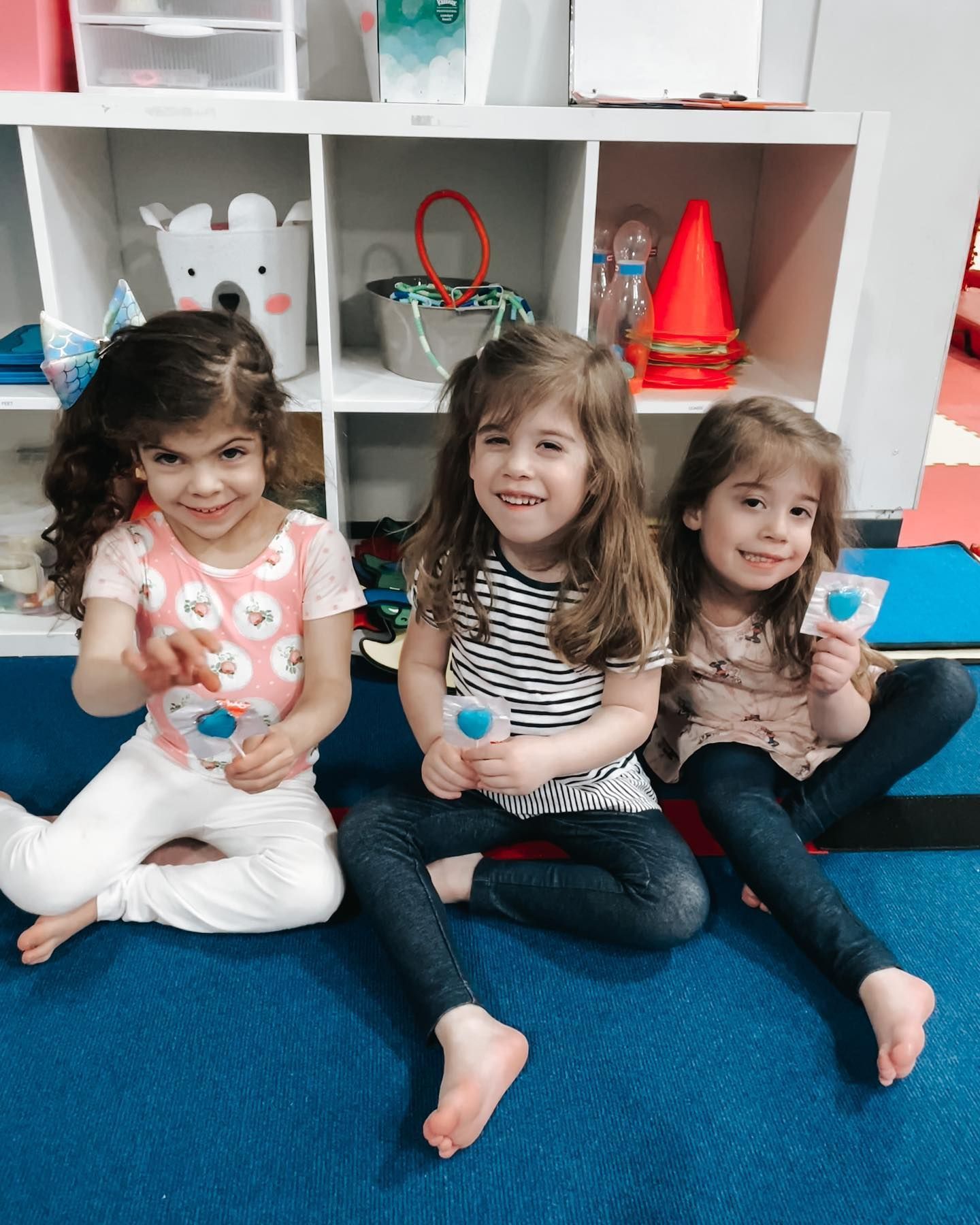 Three children sitting on a blue carpet in front of cubby shelves, smiling and playing with toys.