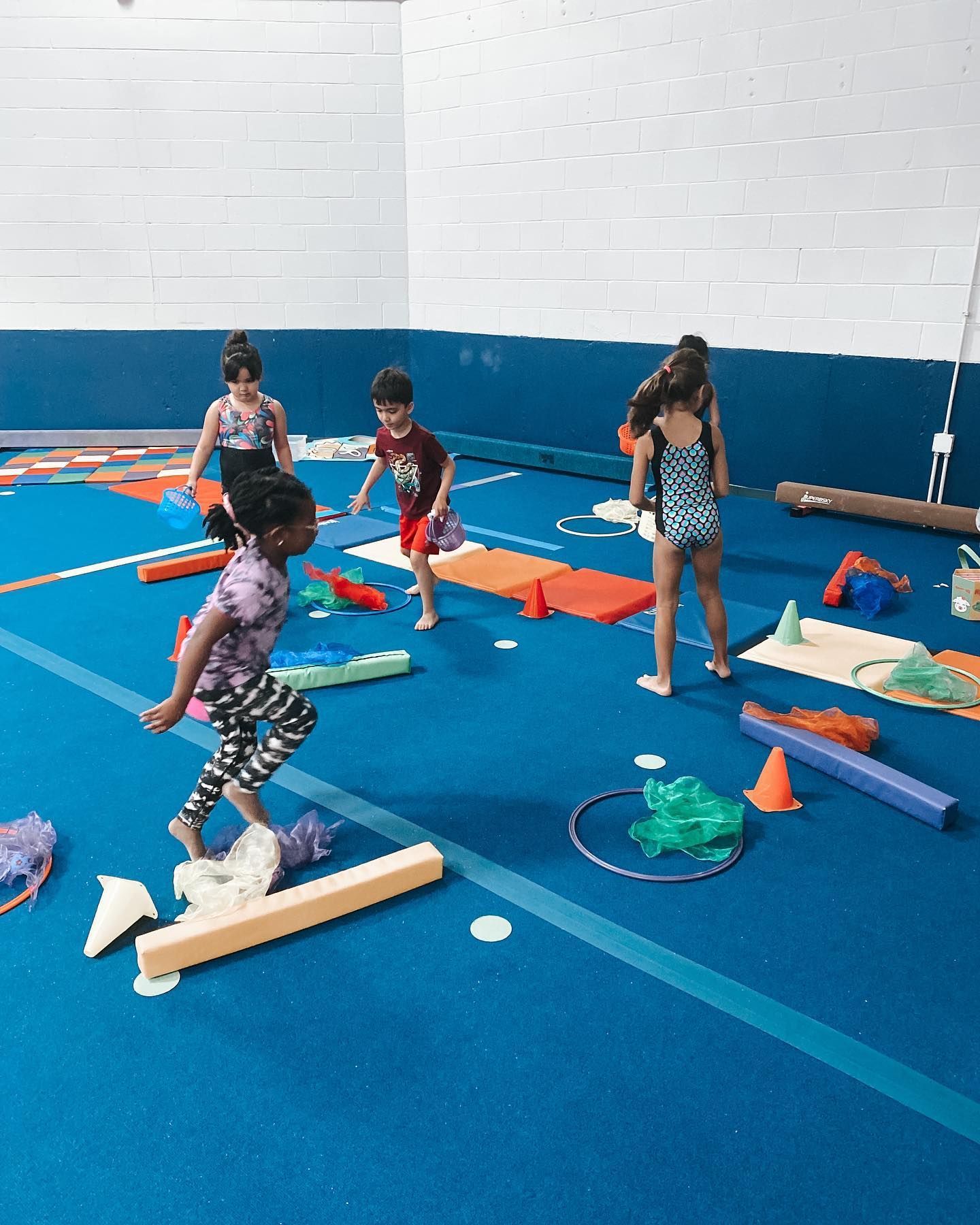 Children navigating a colorful obstacle course in a gym, balancing on beams and stepping over cones.