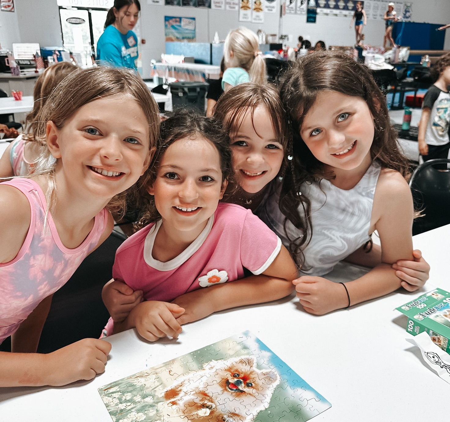 Four smiling children lean over a table with art materials in a classroom.