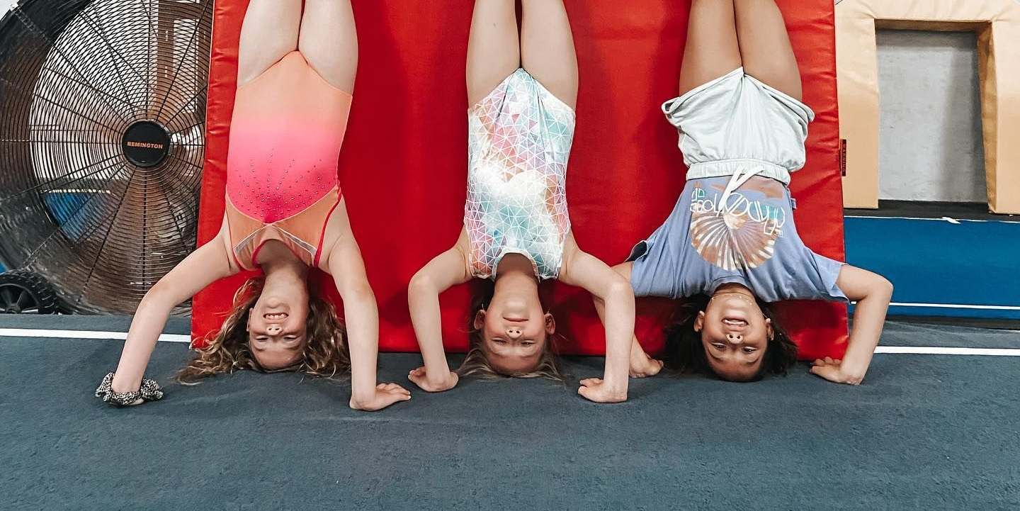 Three children doing handstands outdoors on a mat, smiling in colorful athletic outfits
