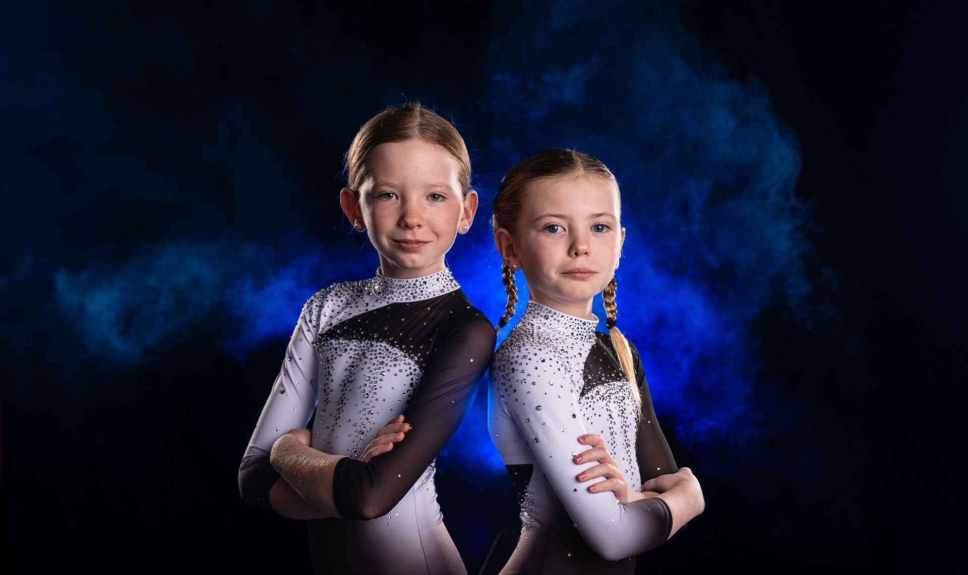 Two figure skaters in white-and-black costumes posed back-to-back against a blue smoky backdrop