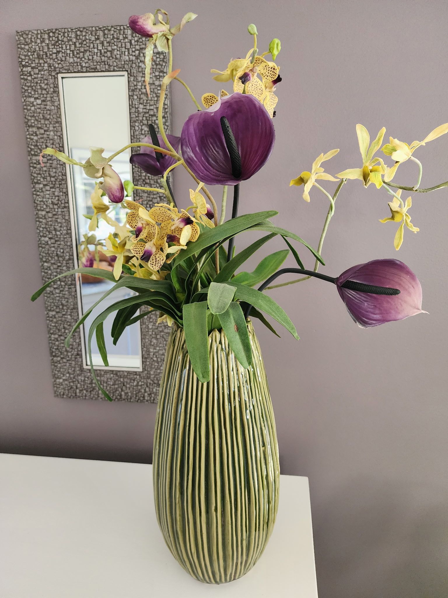 A vase with purple and yellow flowers sits on a white surface near a textured mirror.