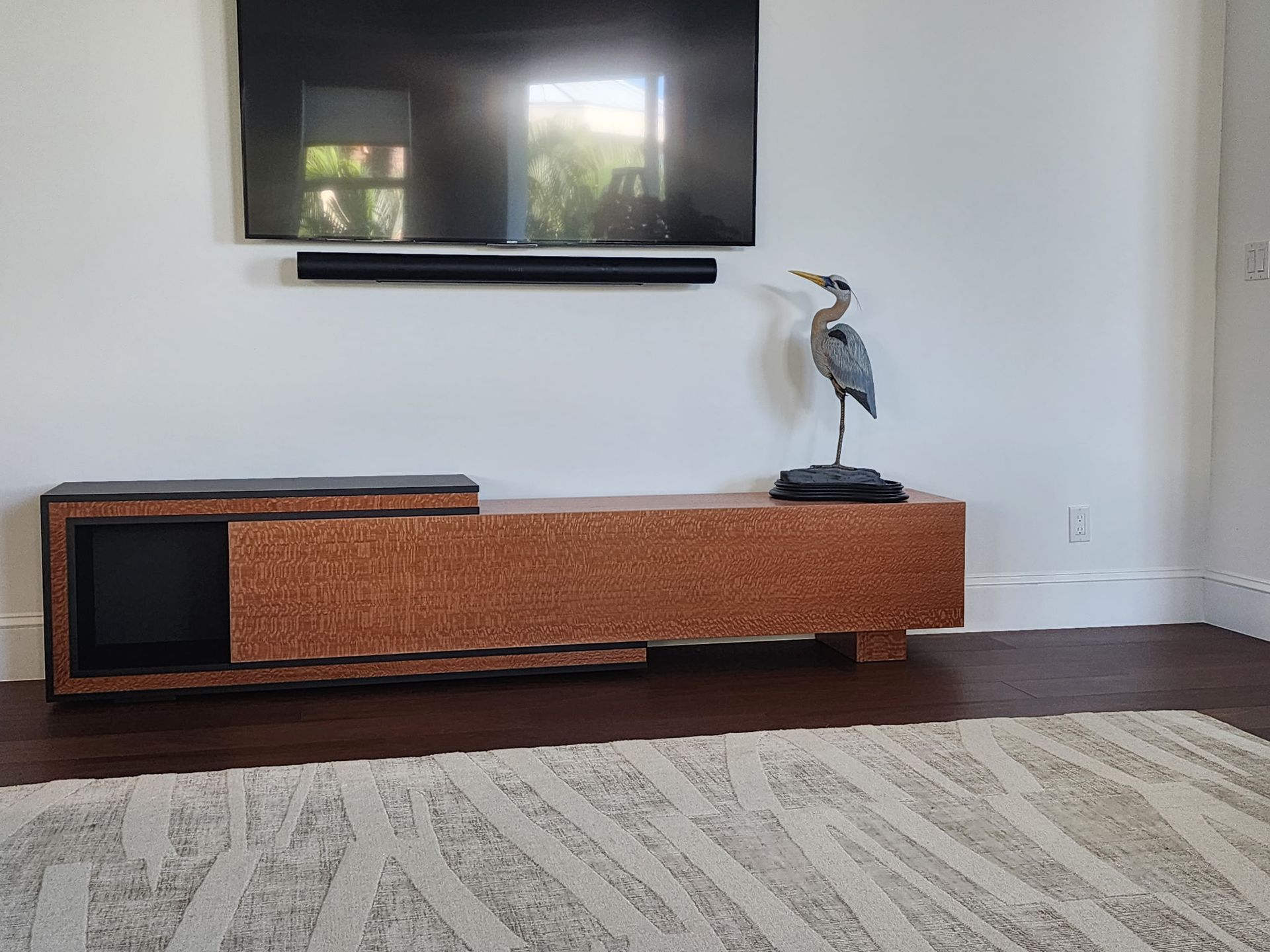 A copper-textured TV console sits under a wall-mounted TV, with a bird sculpture on top and a patterned rug in front.