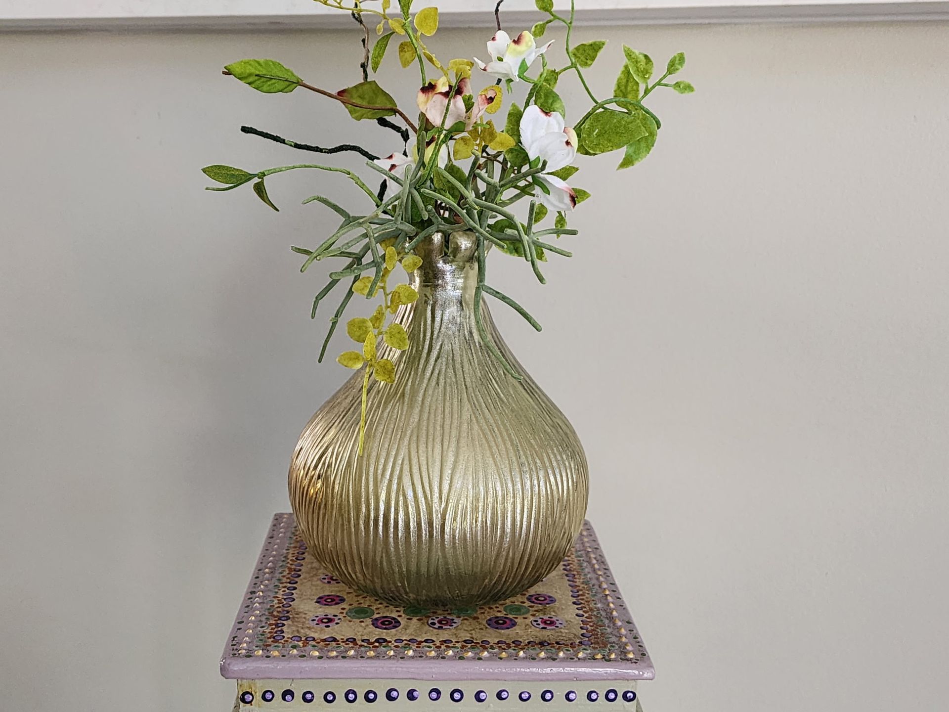 Gold textured vase with floral arrangement on a decorative, tiled stand.