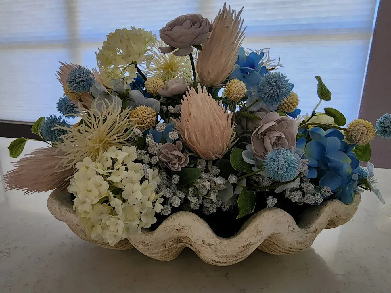 Floral arrangement in a clam-shaped container, featuring blue, yellow, and beige flowers.