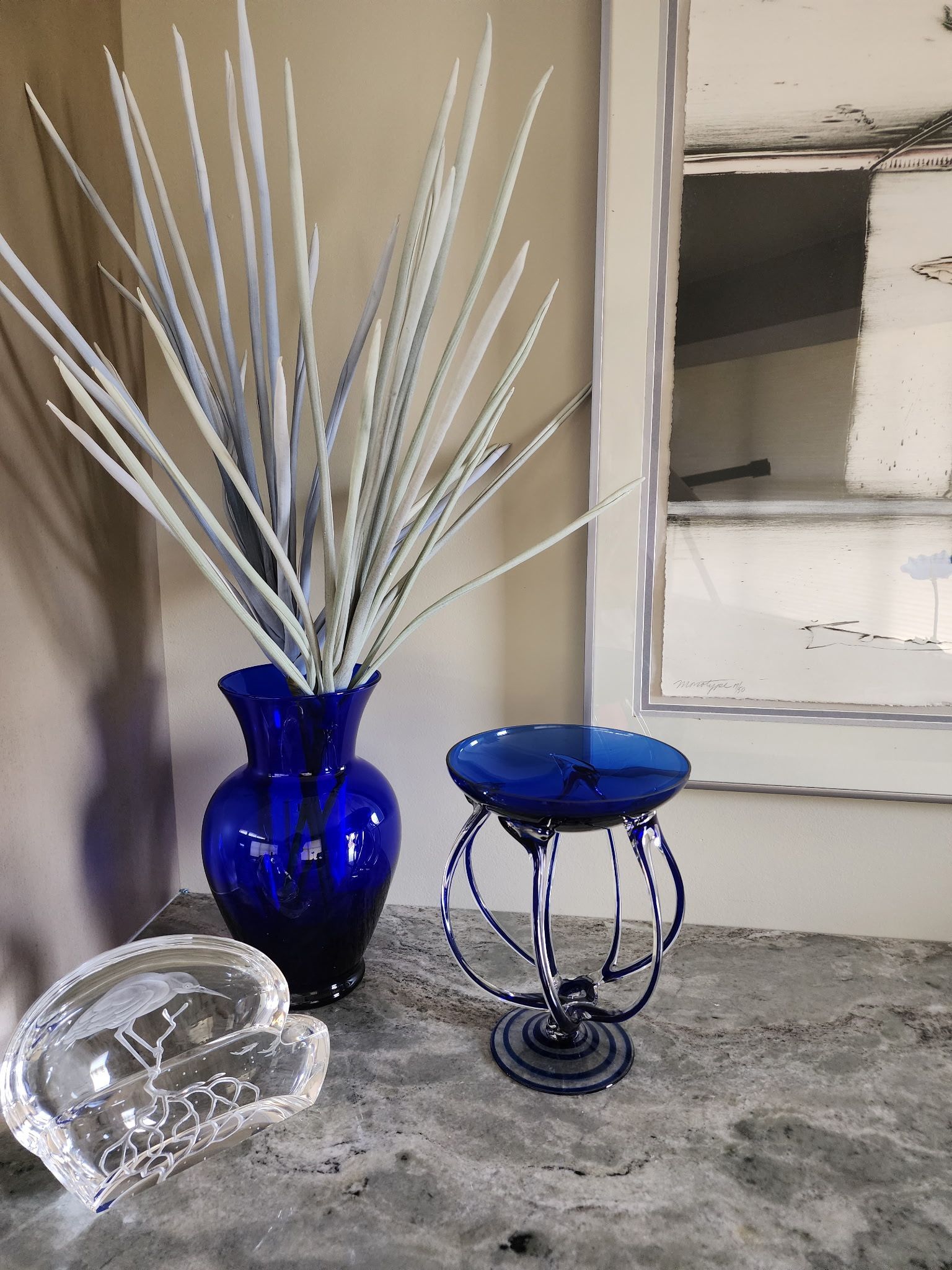 A cobalt blue vase with tall white stems sits beside a blue glass pedestal bowl and a clear crystal piece on a counter.