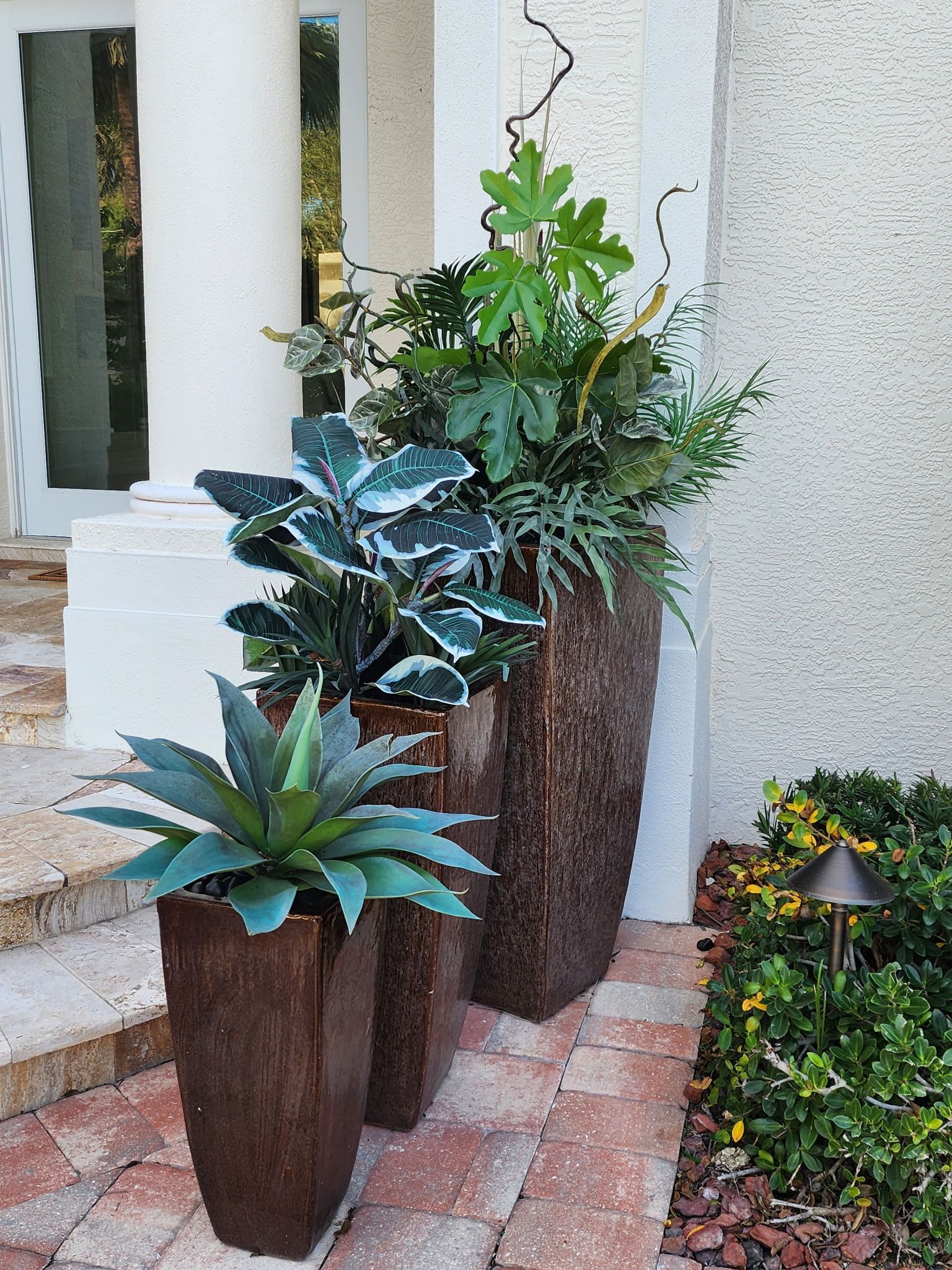 Three tall, brown, textured planters holding different green plants sit on a brick walkway near a white column and wall.