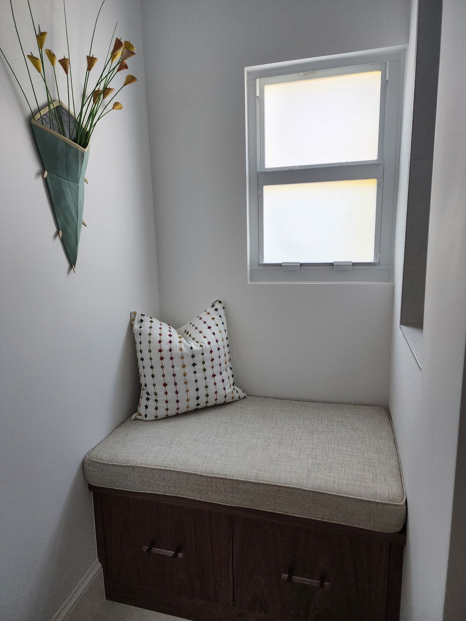 A cozy nook with a brown storage bench, a patterned cushion, and a wall-mounted planter with flowers beneath a window.