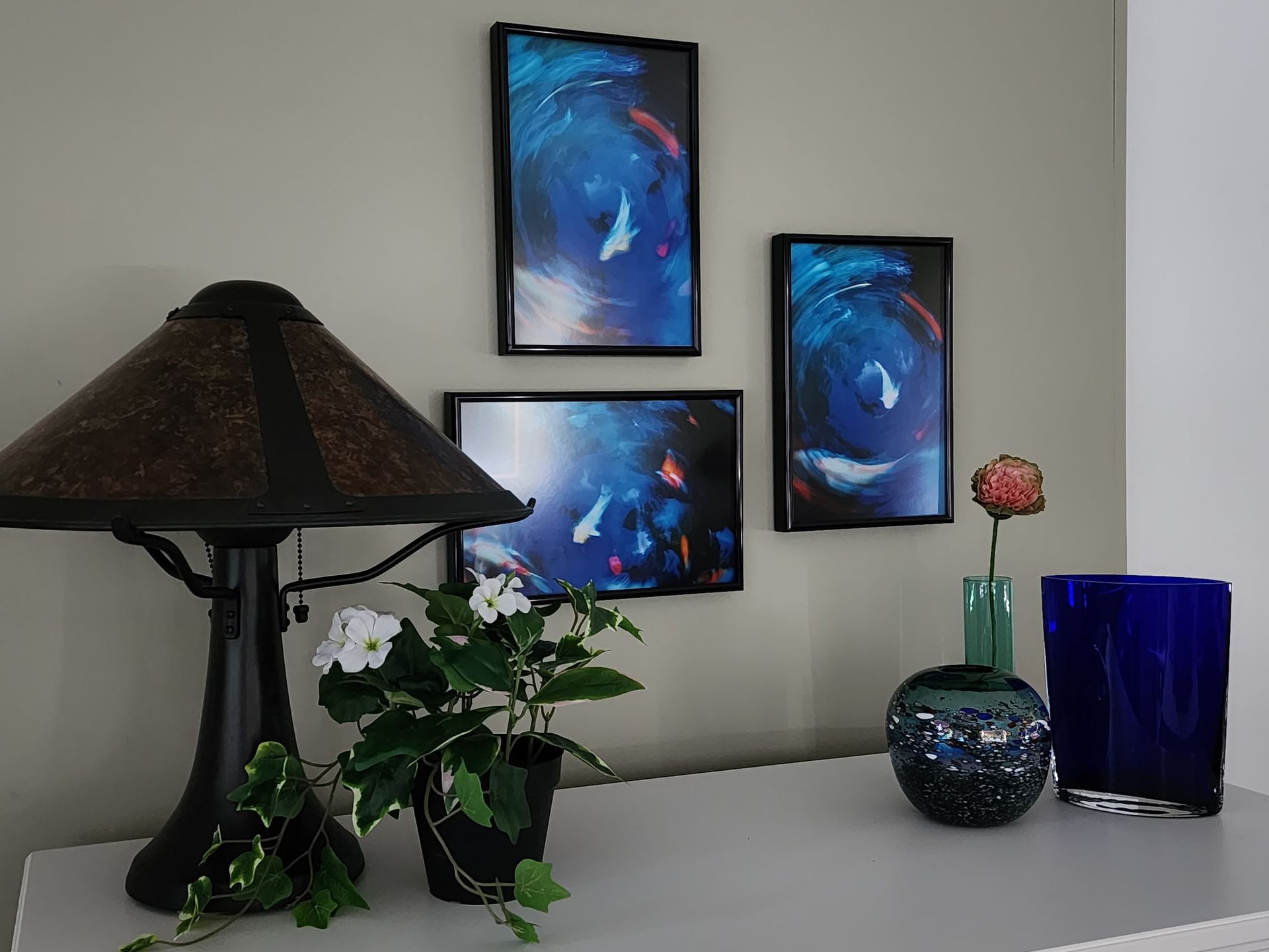 A table holding a dark lamp, a white-flowered plant, a flower in a vase, and a blue vase, with three framed art prints above.