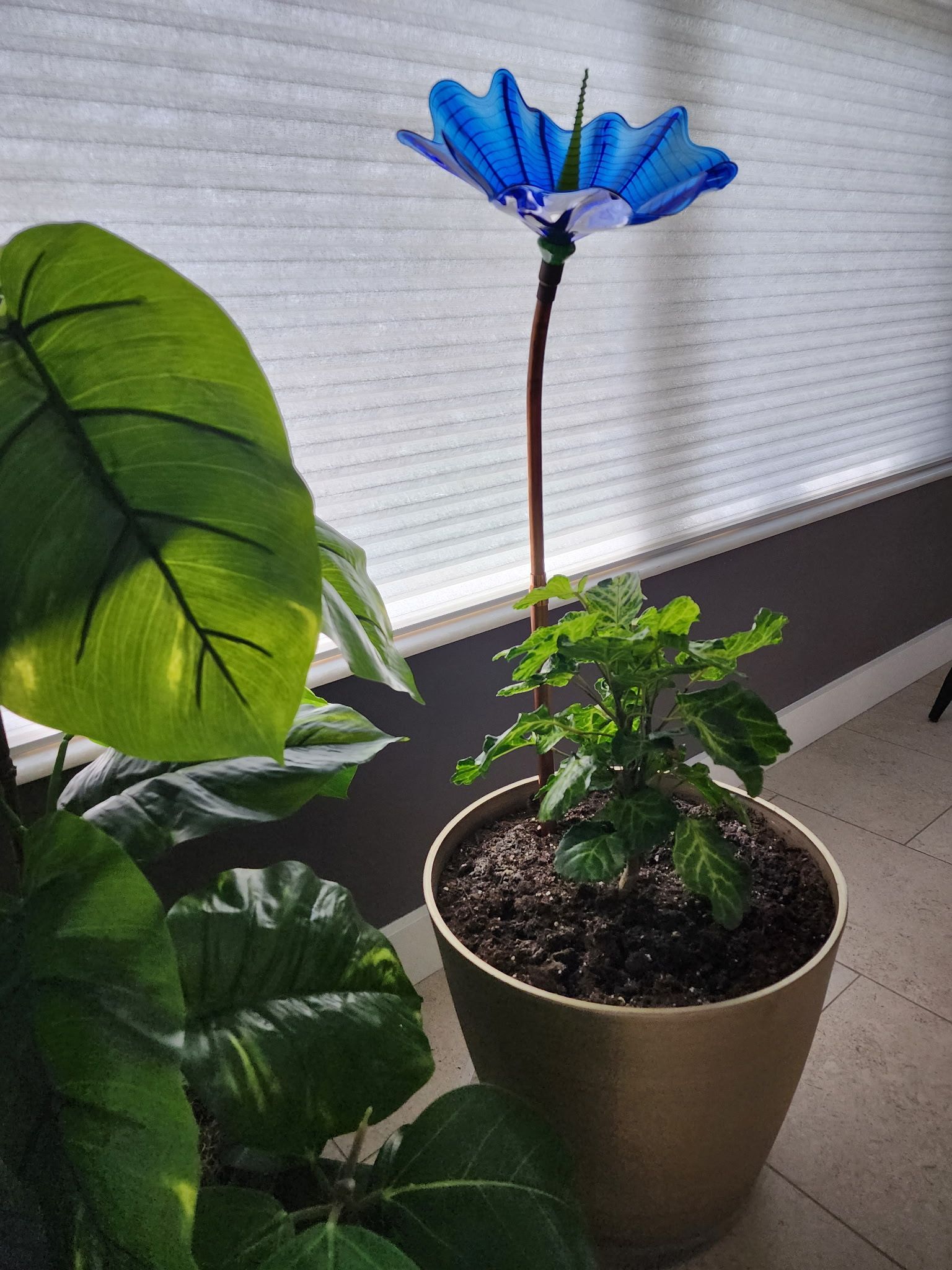 A tall, blue decorative glass flower sculpture stands in a potted plant on a bright indoor windowsill.