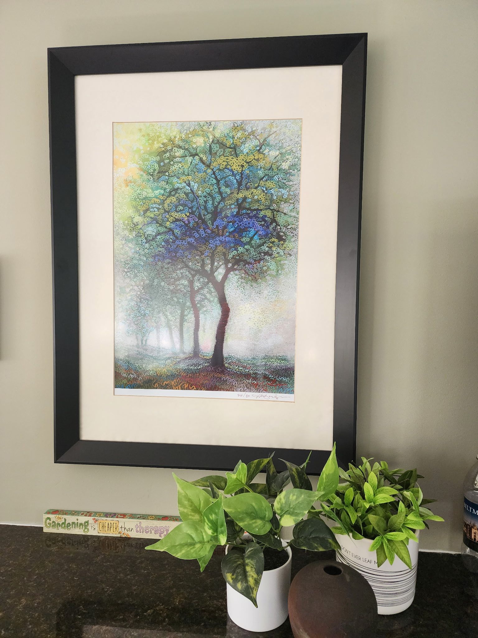 A framed print of a colorful, impressionist-style tree hanging on a wall above two small potted plants.