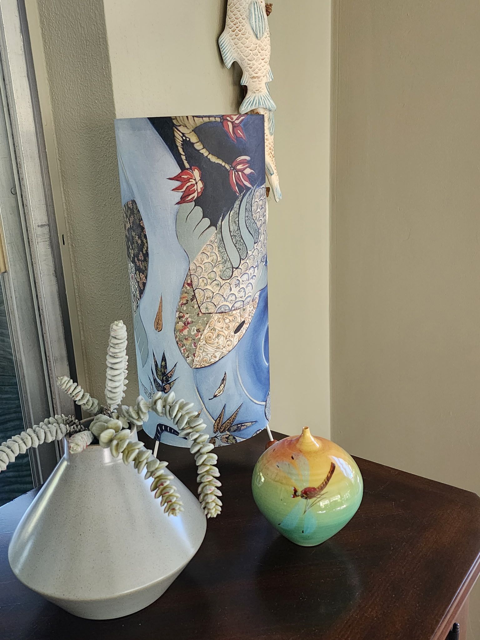 A white vase with a succulent, a colorful glass sphere, and a tall floral wall hanging sitting on a wooden surface.