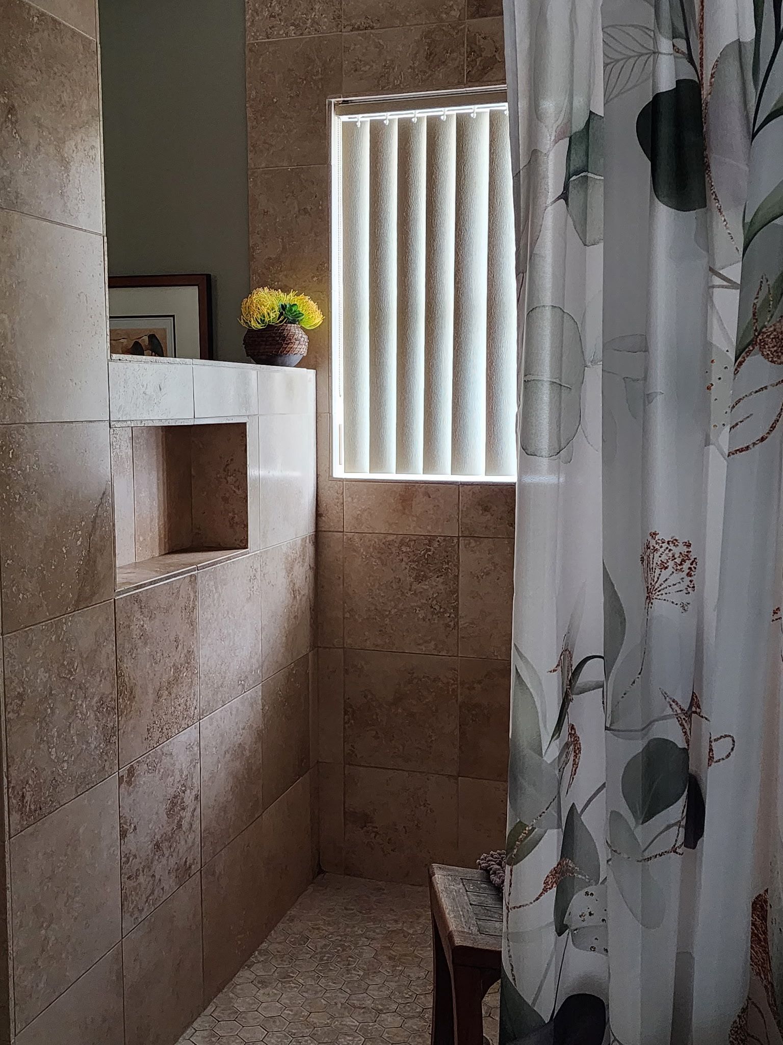 A bathroom shower area with stone-tiled walls, a floral curtain, a small recessed shelf, and a window with white blinds.