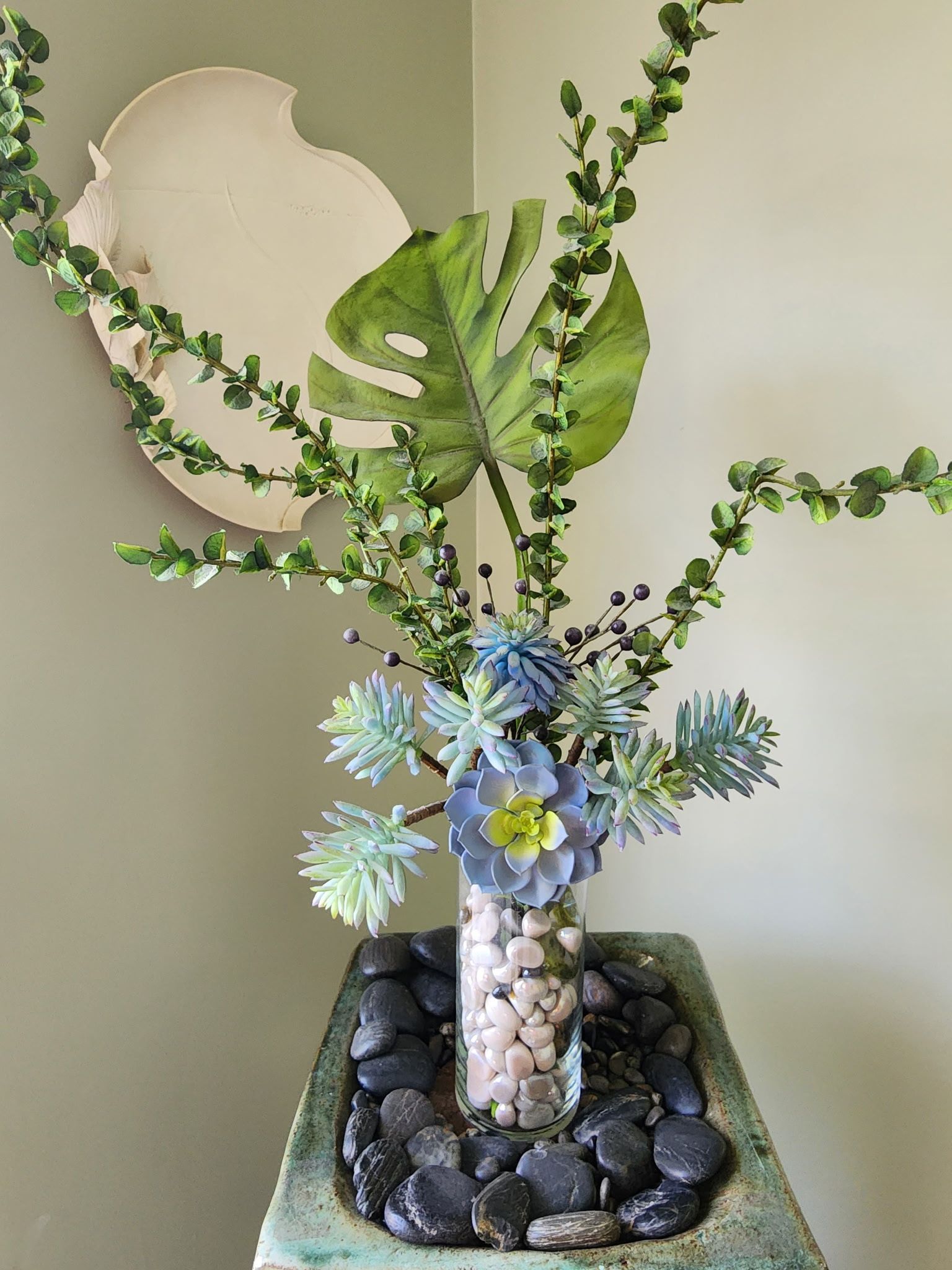 Floral arrangement in a glass vase with small rocks, set on a stone container, with a decorative wall piece in the background.