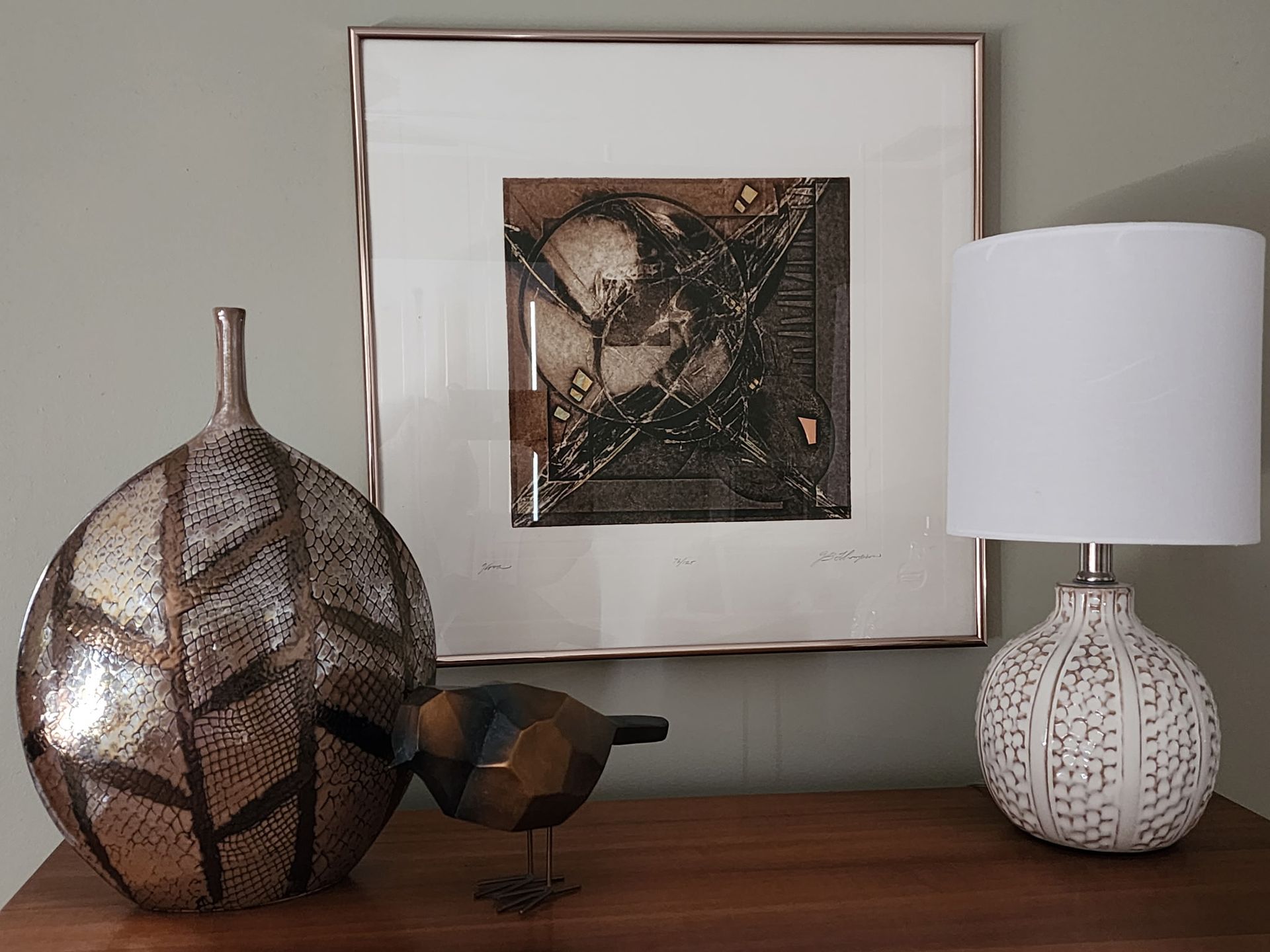 A wooden table with a silver vase, bird sculpture, framed art, and a white lamp against a gray wall.