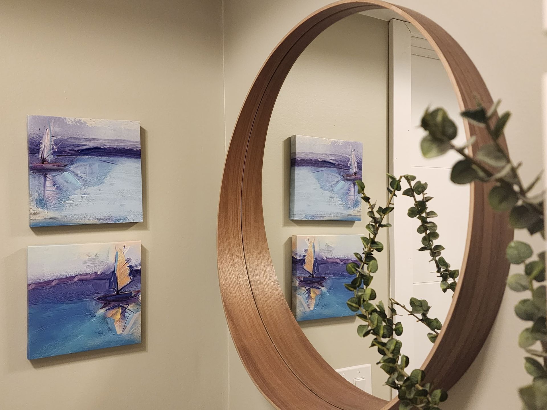 Two abstract ocean paintings on a wall near a round mirror, reflected in the mirror.