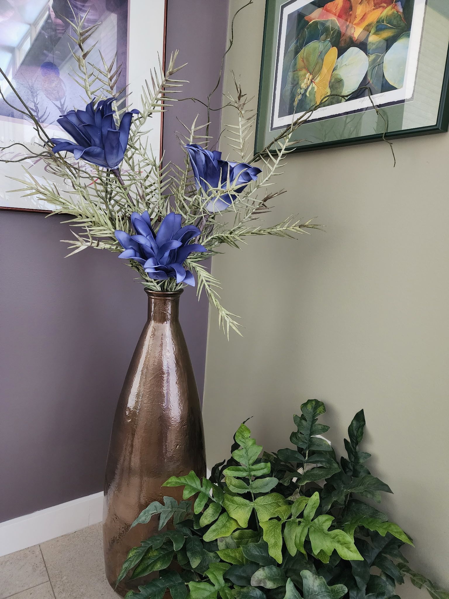 Tall brown vase with blue flowers, artificial greenery in front, paintings on wall.