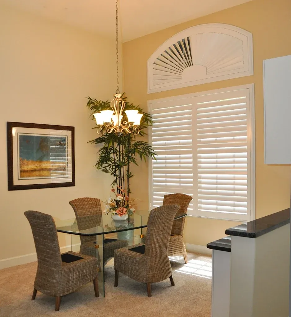Dining room with glass table, woven chairs, chandelier, shutters, art, and plant.