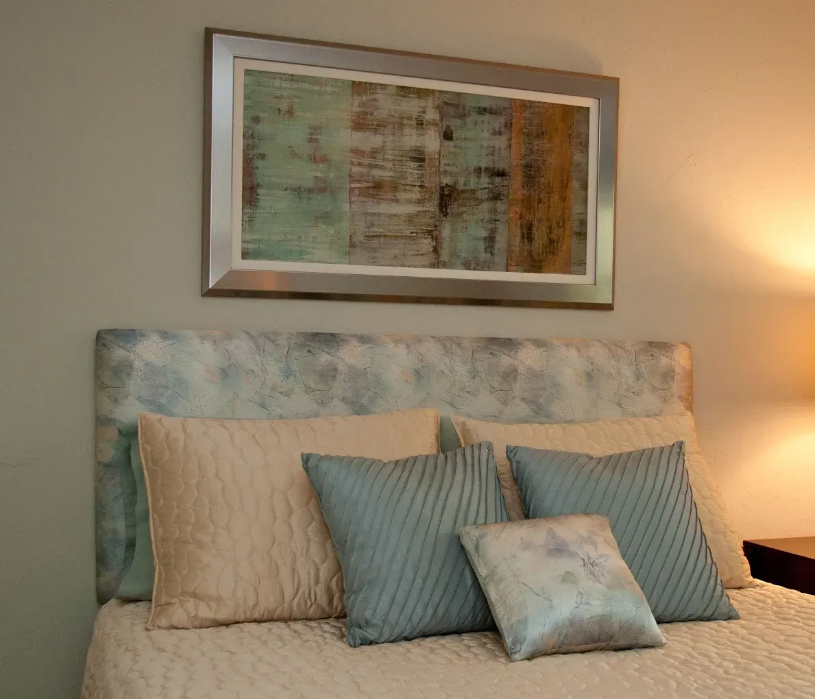 Bed with headboard and pillows, painting above. Beige, blue and brown tones in a bedroom setting.