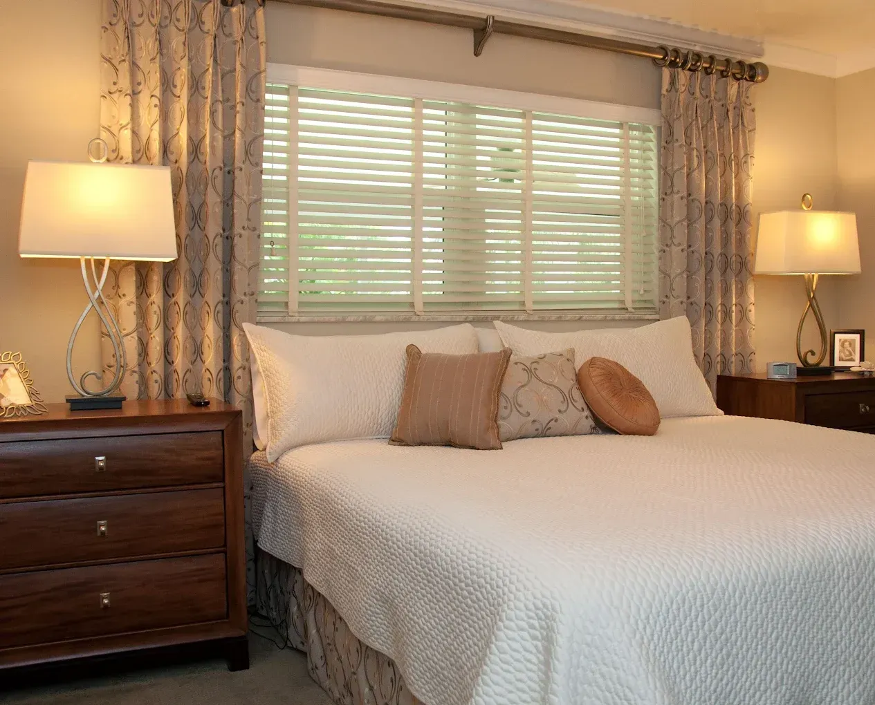 Bedroom with a bed, nightstands, lamps, window with blinds, and patterned curtains.