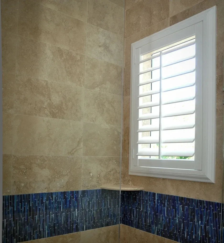 Bathroom with tan tile walls, blue mosaic border, and white shuttered window.