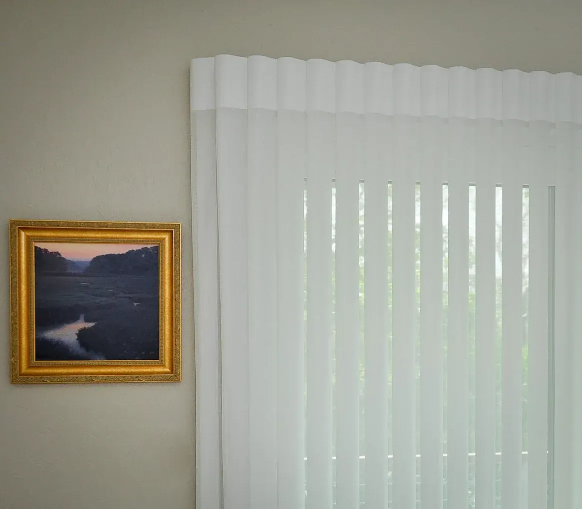 Painting in a gold frame on a light-colored wall next to white vertical blinds.