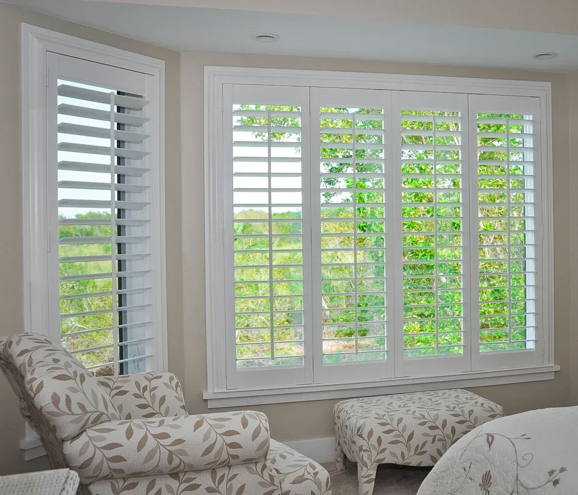 White shutters on a bay window with a view of greenery, and a floral armchair and ottoman.