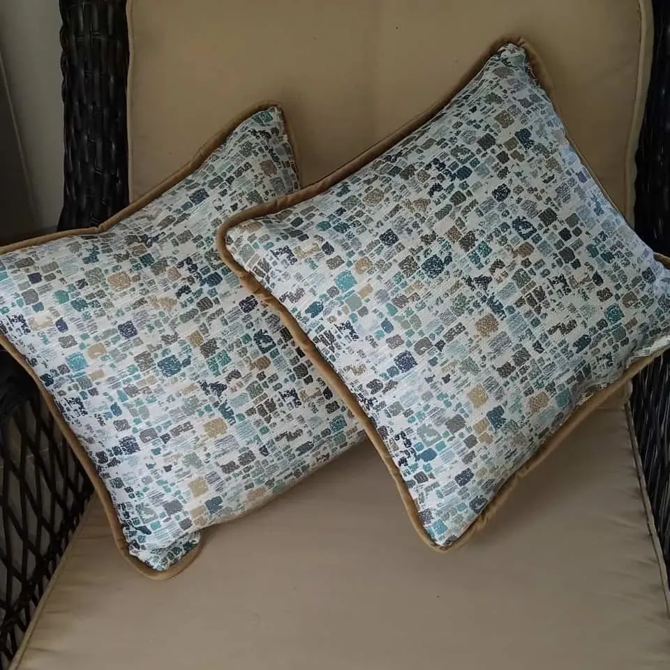 Two square pillows with blue, gray, and tan pattern and brown trim on a beige chair.
