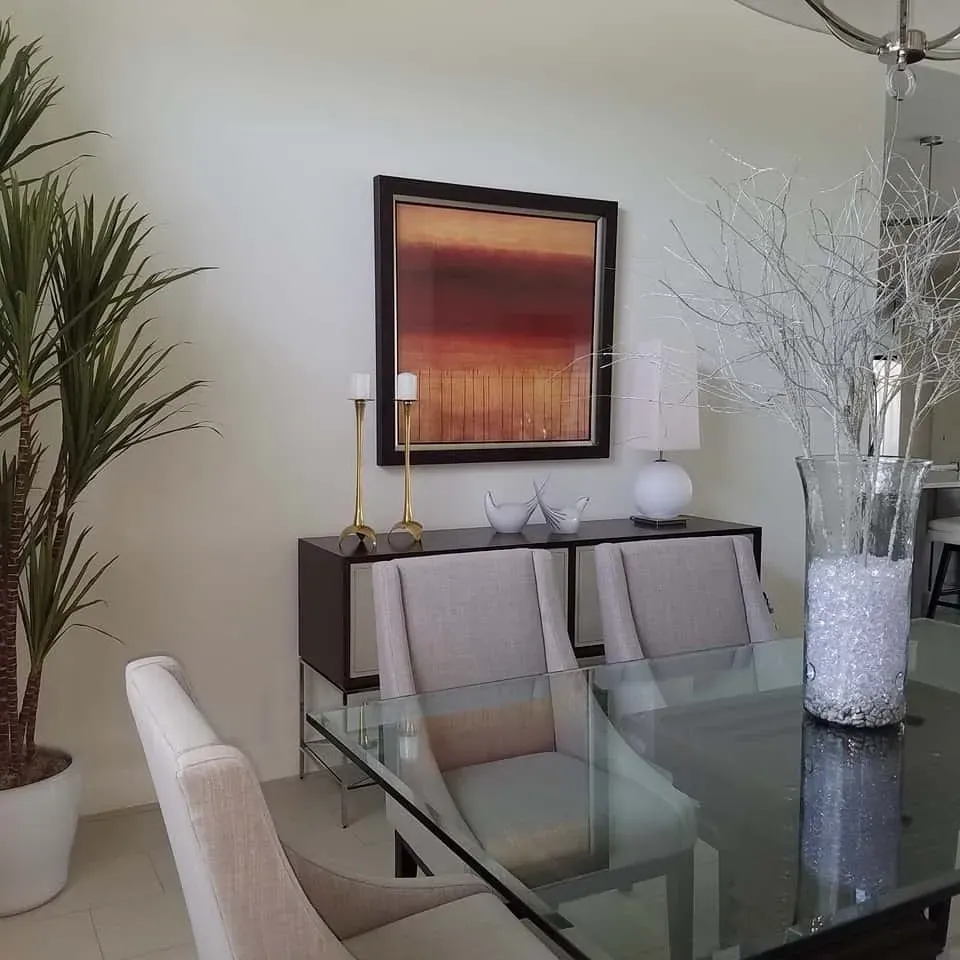 Dining room with glass table, sideboard, artwork, and tall plant.
