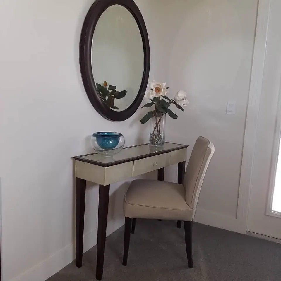 Vanity table with oval mirror, vase of flowers, blue bowl, and chair in a room.