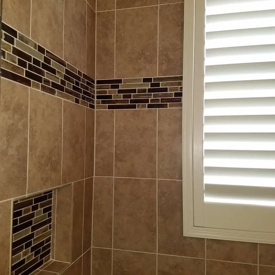 Brown tiled shower with mosaic tile accents and a white shuttered window.