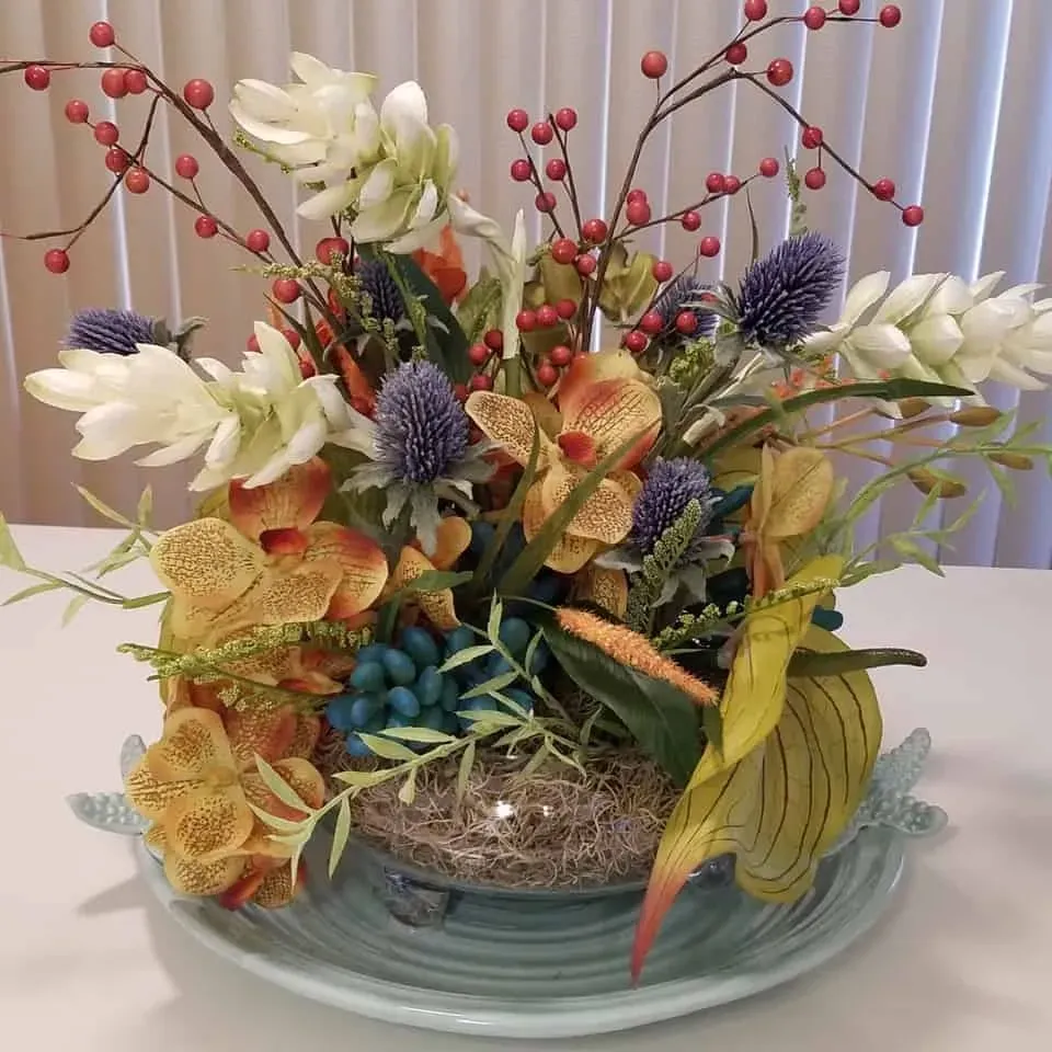 Floral arrangement in a light blue dish, featuring orange, white, and purple flowers with red berries and greenery.