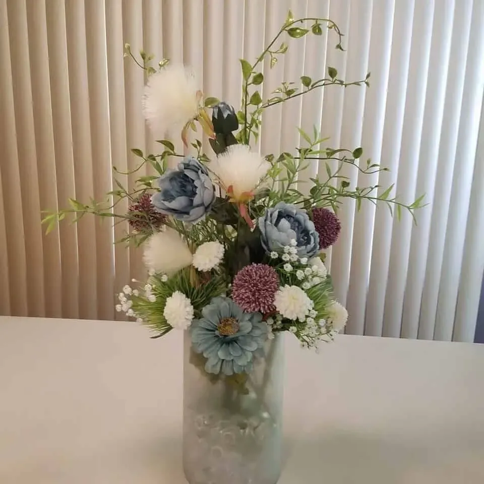 Floral arrangement in a mottled vase; featuring blue, white, and burgundy blooms, with green foliage, in front of a window.