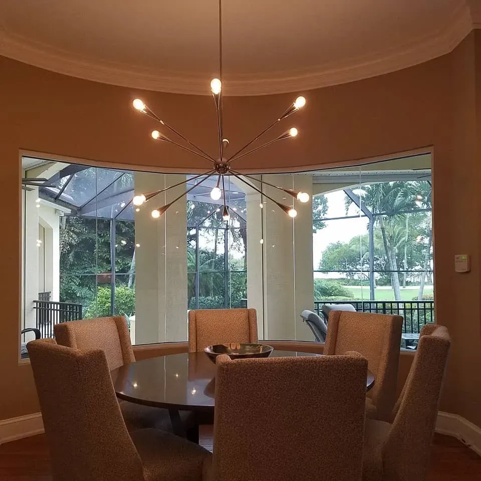Dining room with round table and chairs under a sputnik chandelier, in front of a large window.